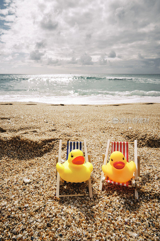 在一个晴朗的多云/阳光明媚的日子里，两只橡皮鸭坐在康沃尔波思科诺海滩的迷你躺椅上。
